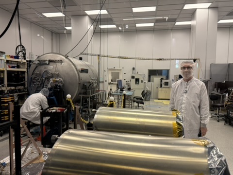 Craig Hardgrove in a white cleanroom bunny suit next to the thermal-shrouded flight GRNS hardware, with the TVAC chamber visible behind